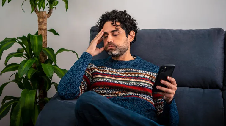 A man on a sofa rubbing his forehead while looking at a misbehaving phone