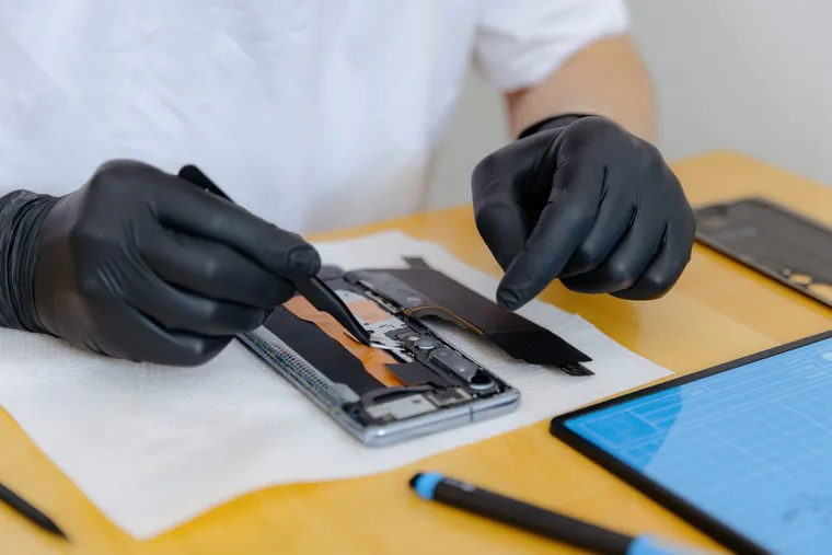 A gloved technician easing a battery out of an opened phone with tweezers