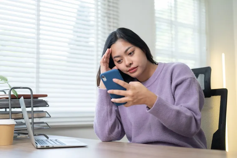 A woman at a desk frowning at her phone beside a laptop